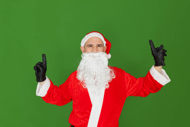 An adult Caucasian man dressed as Santa Claus is dancing with his index fingers up. The background is green. - Photo, Image