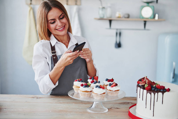 Mujer se para en la cocina y toma una foto de sus galletas caseras y pastel utilizando el teléfono. - Foto, Imagen