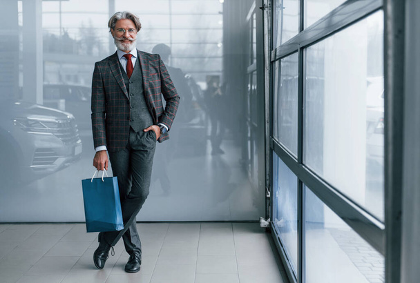 Starszy biznesmen w garniturze i krawacie z siwymi włosami i brodą stojący wewnątrz w niebieskim opakowaniu. - Zdjęcie, obraz