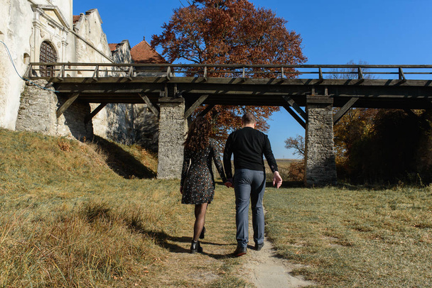 Ein junges Paar läuft Händchen haltend zu uns zurück, im Herbst oder Sommerpark in der Nähe des Schlosses bei Sonnenuntergang. Großaufnahme eines liebenden Paares, das sich beim Gehen an den Händen hält. Selektiver Fokus - Foto, Bild