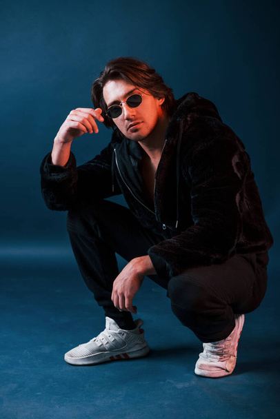 Beautiful man in glasses and black clothes sits in the studio with blue neon lighting. - Valokuva, kuva