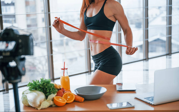 Female vlogger with sportive body standing indoors with measuring tape near table with healthy food. - Photo, image
