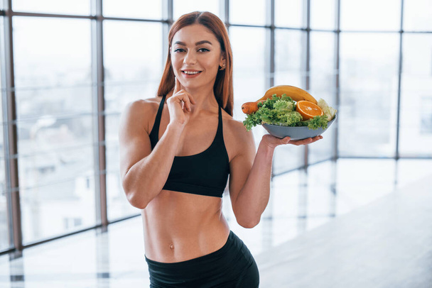Cheerful sportive woman stands indoors with healthy food in hands. - Photo, image