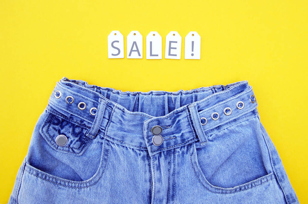 Flat lay, detail of denim jeans with white labels and inscription SALE on yellow background. Shopping and discount concept. Black Friday. - Photo, Image