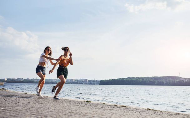 Two female friends runs and have fun at beach near the lake at sunny daytime. - Фото, зображення