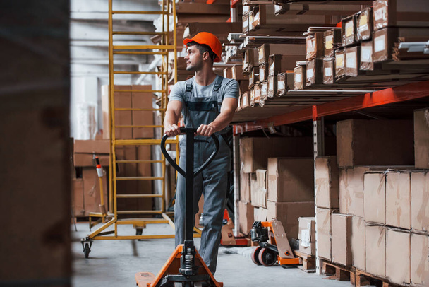 Young male worker in uniform is in the warehouse with pallet truck. - Valokuva, kuva