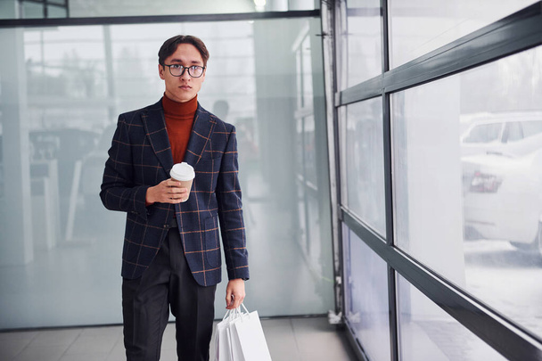 With cup of drink and shopping bags. Young business man in luxury suit and formal clothes is indoors in the office. - Photo, image