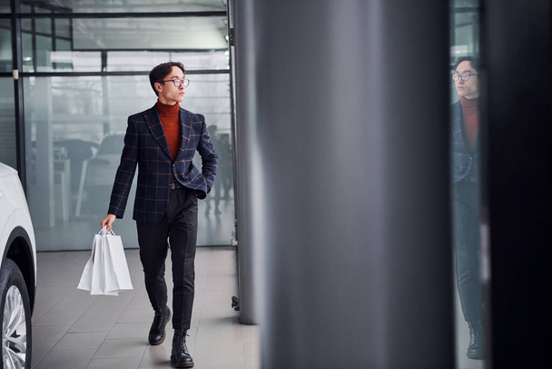 Holding shopping bags. Young business man in luxury suit and formal clothes is indoors near the car. - Foto, afbeelding