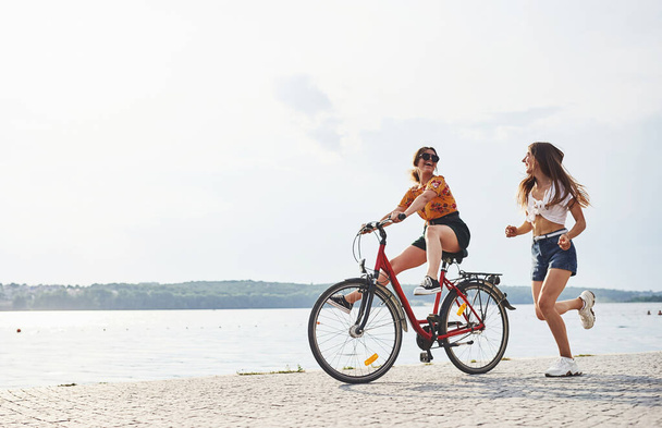 Girl runs near bicycle. Two female friends on the bike have fun at beach near the lake. - Photo, Image