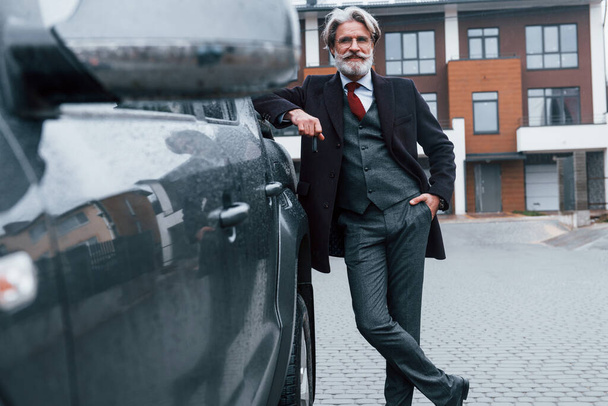 Fashionable senior man with gray hair and beard leaning on his car and holding keys. - 写真・画像