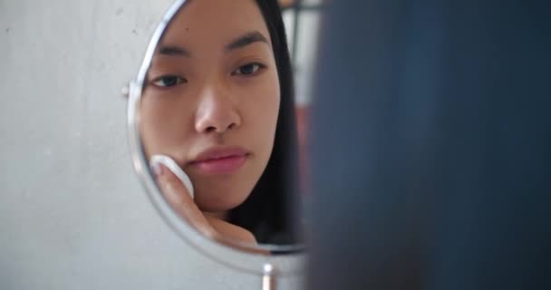 Nette schöne natürliche brünette asiatische Mädchen Putzgesicht mit Baumwollschwamm lächelnd ihr Spiegelbild in kleinen Kosmetikspiegel betrachtend. Attraktive Frau entfernt Make-up mit Reinigungsmittel und Wattepads - Filmmaterial, Video
