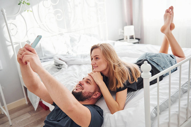 Young married couple making a selfie by using phone in bedroom at daytime. - Fotoğraf, Görsel