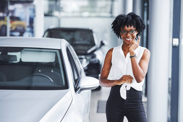 Young african american woman in glasses stands in the car salon near vehicle. - Zdjęcie, obraz