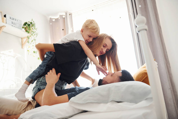 Young married couple with their young son lying down on bed and have fun together in bedroom at daytime. - Φωτογραφία, εικόνα