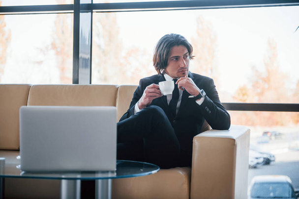 Portrait of handsome young businessman in black suit and tie that sits on the sofa and takes a break. - 写真・画像