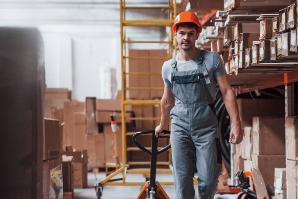 Young male worker in uniform is in the warehouse with pallet truck. - 写真・画像