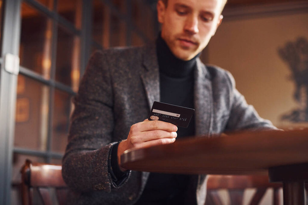 Portrait of modern young guy in formal clothes that sits in the cafe and holds credit card in hand. - Foto, immagini