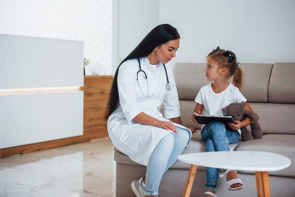 Vrouwelijke arts met notitieblok zit in de kliniek met een klein meisje. - Foto, afbeelding