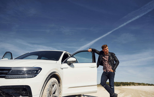 Man in black leather jacket stands near his parked white car outdoors against blue sky. - Foto, Imagem