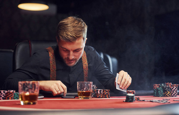Elegant young man sits in casino, using phone and plays poker game. - 写真・画像