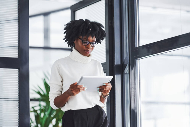 Young african american woman in glasses stands indoors in the office with tablet in hands. - 写真・画像