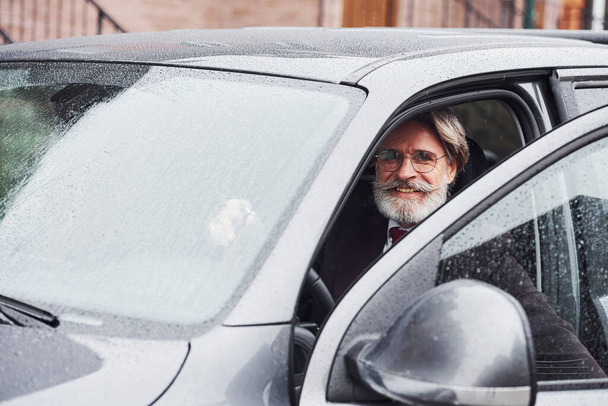 Fashionable senior man with gray hair and beard is outdoors on the street sitting in his car. - Zdjęcie, obraz