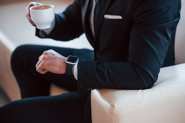 Close up view of young businessman in black suit and tie with cup f drink checking time. - Foto, Imagem