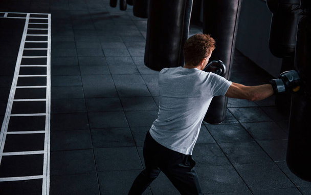 Young man in white shirt and boxing protective gloves doing exercises in gym with pushing bag. - Φωτογραφία, εικόνα