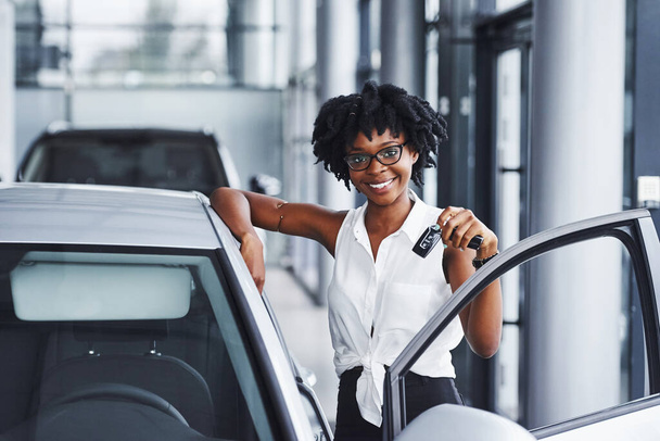 Young african american woman in glasses stands in the car salon near vehicle with keys in hands. - Fotó, kép