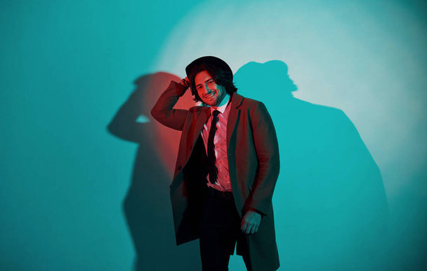Portrait of young stylish man in hat, suit and tie that stands in neon lights in the studio. - Фото, изображение