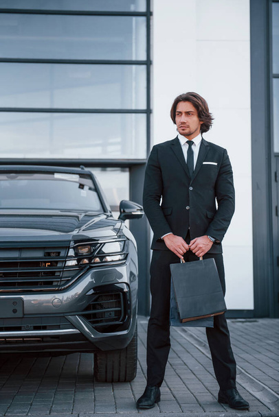 Portrait of handsome young businessman in black suit and tie outdoors near modern car and with shopping bags. - Photo, image