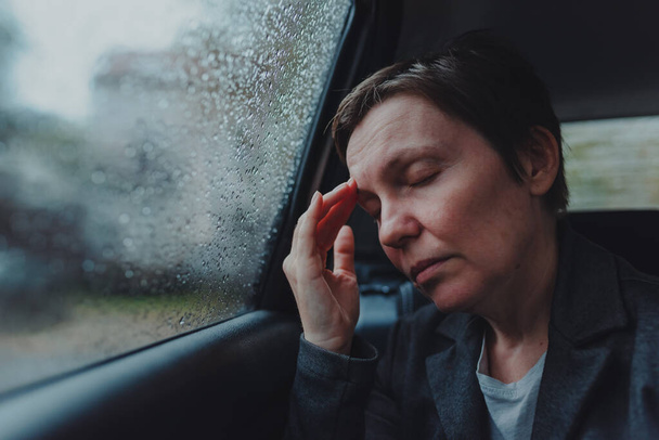 Zakenvrouw met ernstige hoofdpijn zittend op de achterbank van een auto tijdens regen, selectieve focus - Foto, afbeelding