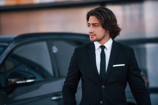 Portrait of handsome young businessman in black suit and tie outdoors near modern car. - Foto, afbeelding