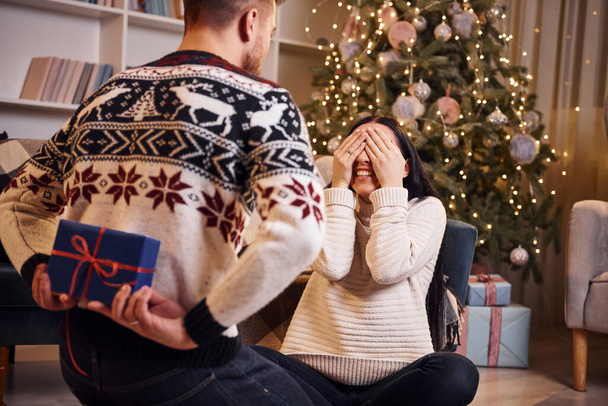 Man covers eyes of hir girlfriend and giving new year surprise in christmas decorated room. - Foto, immagini