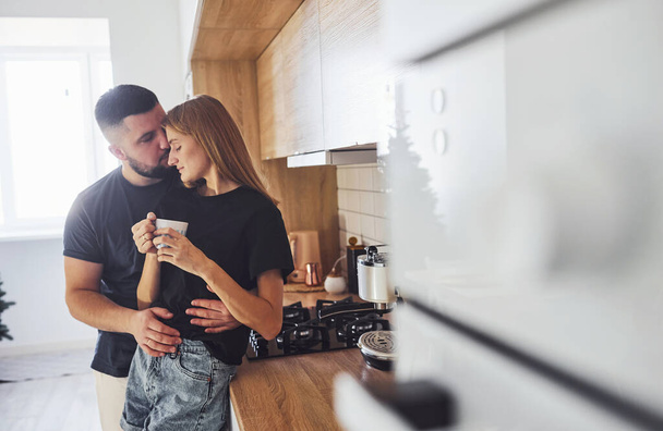 With cup of fresh drink. Morning for young married couple that standing indoors in the kitchen. - Фото, зображення