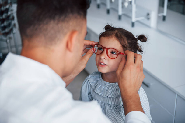 Молодий педіатр у білому пальто допомагає отримати нові окуляри для маленької дівчинки
. - Фото, зображення