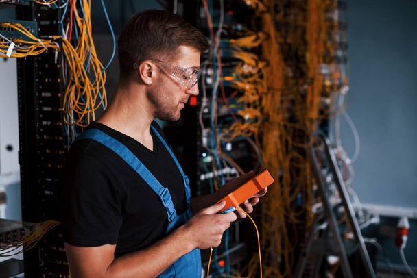 Young man in uniform with measuring device works with internet equipment and wires in server room. - 写真・画像