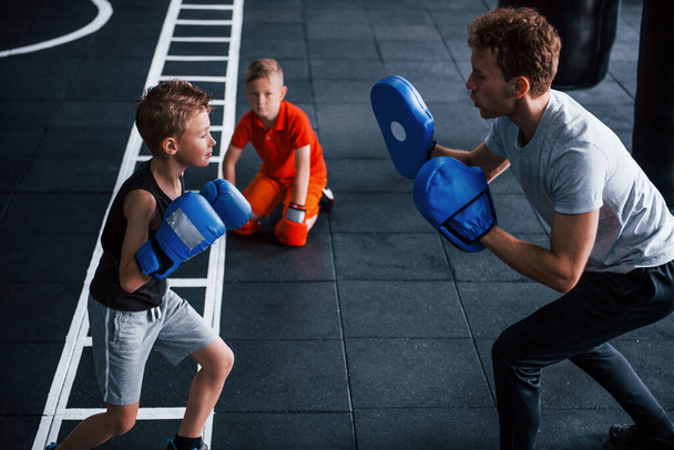Young trainer teaches kids boxing sport in the gym. - Фото, изображение