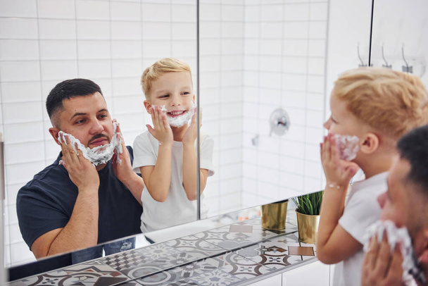 Father with his son is in the bathroom have fun by using shaving gel and looking in the mirror. - Foto, Imagem