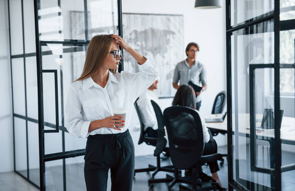 Brunette vrouw in bril met kopje drank in handen staat in het kantoor tegenover medewerkers. - Foto, afbeelding