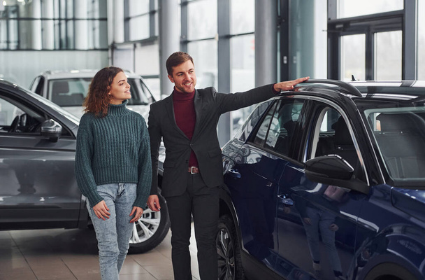 Επαγγελματίας πωλητής βοηθώντας νεαρή κοπέλα επιλέγοντας νέο σύγχρονο αυτοκίνητο σε εσωτερικούς χώρους. - Φωτογραφία, εικόνα
