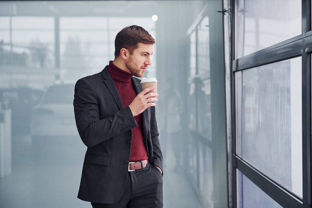 Young business man in luxury suit and formal clothes wth cup of drink is indoors in the office. - Photo, Image