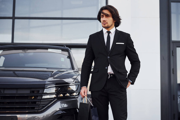 Portrait of handsome young businessman in black suit and tie outdoors near modern car and with shopping bags. - Foto, immagini