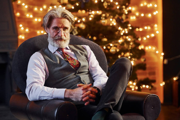 Portrait of stylish senior with grey hair and beard is in decorated christmas room sitting on the chair. - Foto, immagini