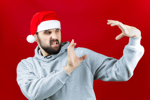 egy szakállas férfi karácsonyi ruhákban, piros háttérrel utánozza az ajándékot a kezében. - Fotó, kép