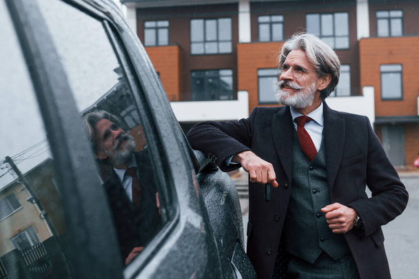 Fashionable senior man with gray hair and beard leaning on his car and holding keys. - Φωτογραφία, εικόνα
