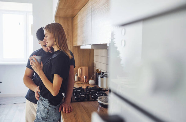 With cup of fresh drink. Morning for young married couple that standing indoors in the kitchen. - Foto, afbeelding