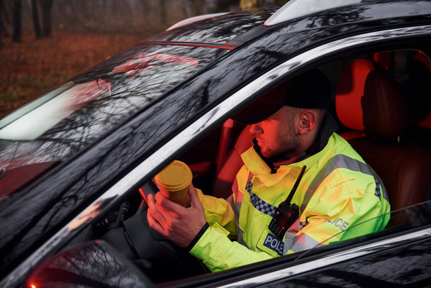 Male police officer in green uniform sitting in automobile with cup of drink. - Foto, Imagen