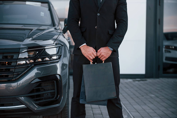 Close up view of businessman in black suit and tie stands near modern automobile with shopping bags in hands. - Foto, afbeelding