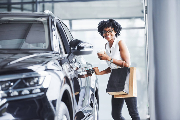 Young african american woman in glasses stands in car salon near vehicle with package in hands. - Foto, Bild